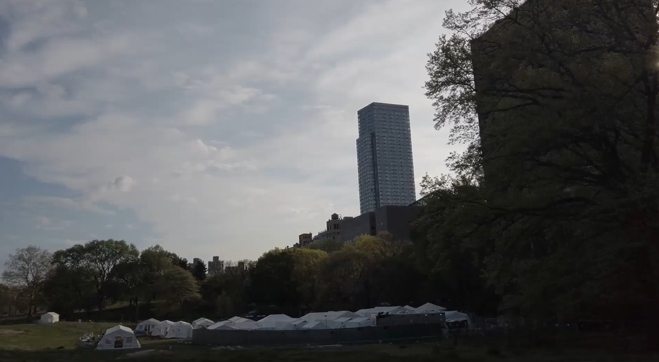 A view of white tents on a lawn, with a city skyline in the background.