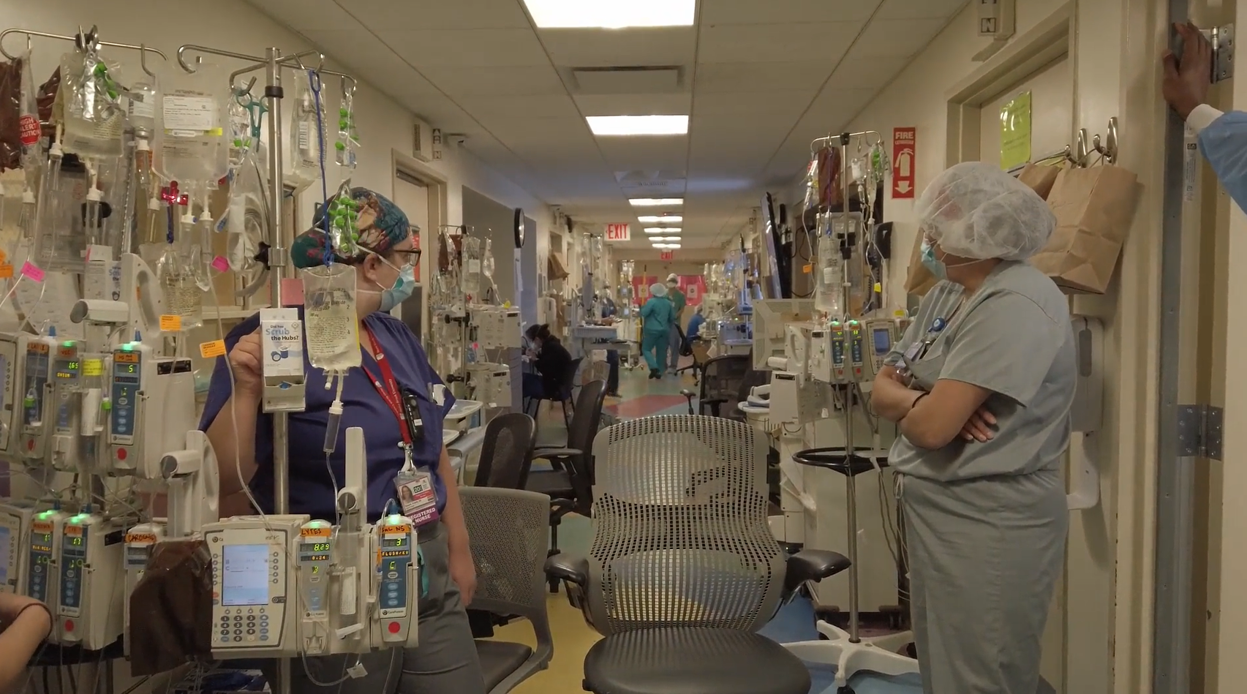 A hospital hallway with a couple people in scrubs and facemasks, crowded with desk chairs and IV carts.