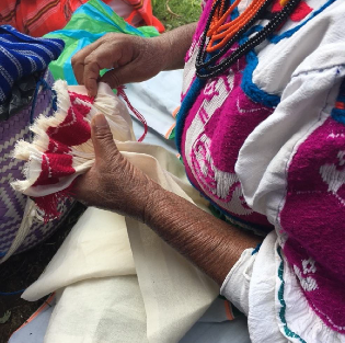 An upclose shot of a woman from San Pablo Tijaltepec embroidering a piece of white fabric with red yarn. The design is similar to the one she wears on her blouse, which is embroidered with pink yarn, and she wears black and orange beaded necklaces. The shot only frames her hands and torso.