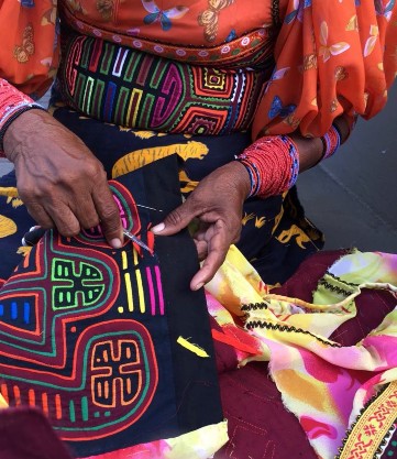 A guna woman shown sewing her mola, framed on her lap, hands and torso. She wears a red blouse with a mola showing green, pink, and yellow lines, on her wrists she wears red beaded bracelets. The mola she sews depicts flowers in green and orange lines, contrasting with the main panel of dark blue fabric.