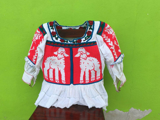 A blouse is propped on a chair in lieu of a manekin, in front of a bright green background. The embroidery depicts on the front the image of sheep, mirrored in the style of embroidery from Tijaltepec. It shows the “negative” effect the embroidery technique creates in these blouses. The sleeves have the image of a rose. The neck is adorned with little embroidered roses.