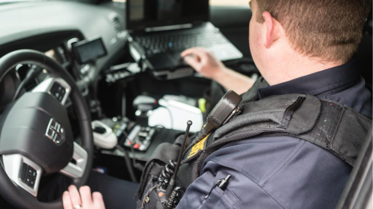 Image of a male police officer sitting in the driver's seat of his vehicle with the door open, looking at a mobile data terminal (police car laptop)