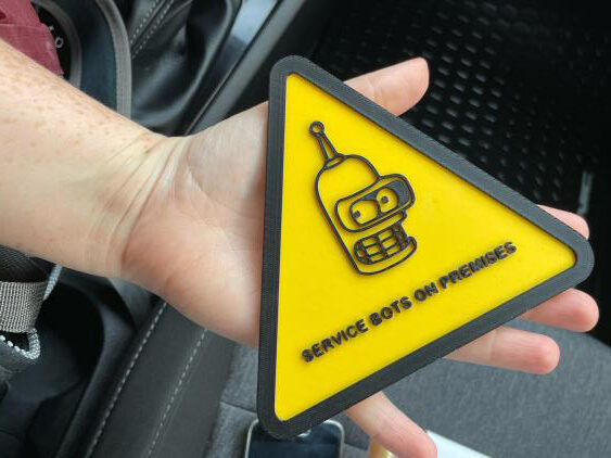 Triangle 3-D printed yellow and black sign reading “Service Bots on Premises” with face of a robot from a popular television show.