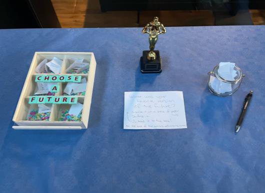 Box labeled “Choose a Future” on display next to an award, and a cup of blank slips of paper for visitors to write their favorite future on and submit to the box.