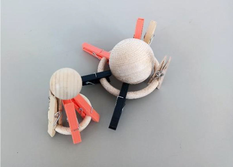 Two wooden rings lie side by side, each with a number of clothespins pinned around them in an array. The clothespins are of mixed colors and materials, some unstained wood and some black or orange plastic. The smaller ring on the left has its clothespins pointed skyward, creating a pinnacle where a small wooden ball rests. The larger ring on the right has its once-skyward clothespins splayed flat, with a large wooden ball resting in the center. The smaller ensemble represents the functional support network for fast-food consumption (on the left side). The larger ensemble represents the dysfunctional support network for healthy eating.