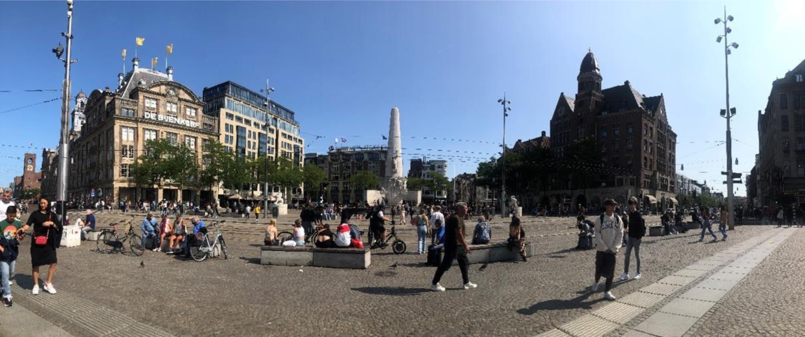 Panorama of Amsterdam's Dam Square monument and pedestrians walking by on the street. There is a bright blue sky and shopping buildings surround the square