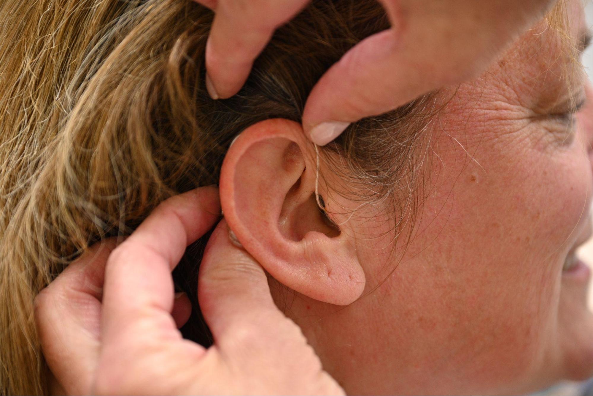 Figure 3. A close-up photograph of a woman's ear. Another person's hands are in frame holding back her hair and fitting the wire and mic of a hearing aid behind her ear.