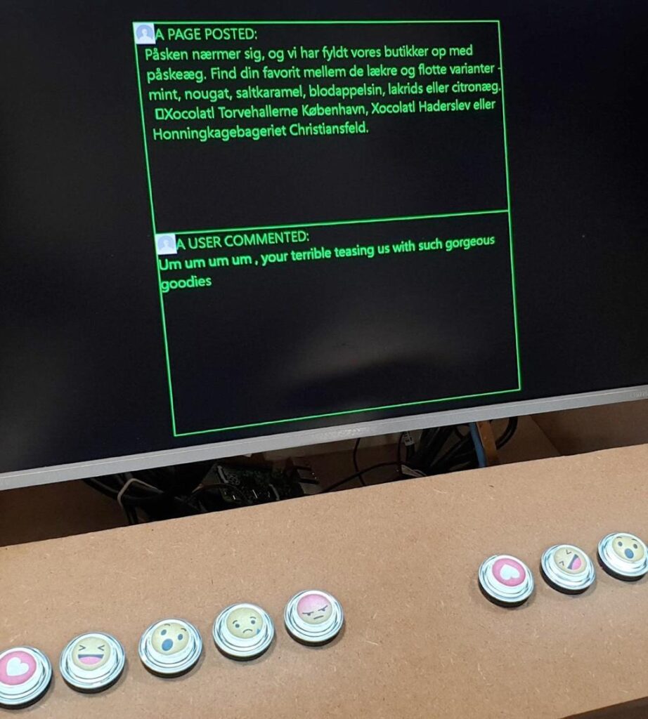 A picture of ‘the thick machine’ which shows the a black screen with green letters showing the post of a Facebook page as well as an example of a user comment on the given post. The screen is attached to a wooden board with button carrying icons of the Facebook emojis ‘heart’. ‘laugh’, ‘wow’, ‘sad’ and ‘angry’. These buttons can be pressed by the person playing the game.