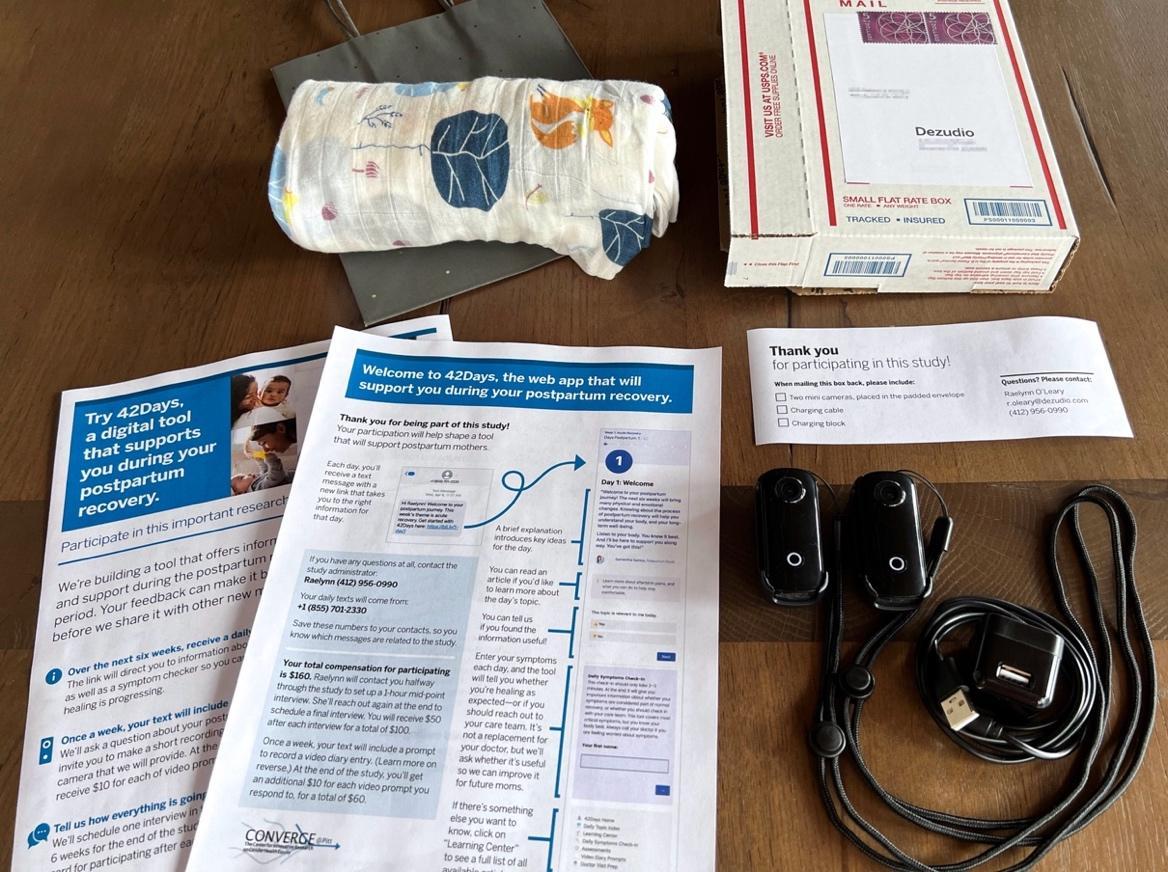 Contents of the study kit arranged on a table. Items include a bag, a baby blanket, study participation instructions, two small portable cameras and a charger, and a stamped addressed mailing box.